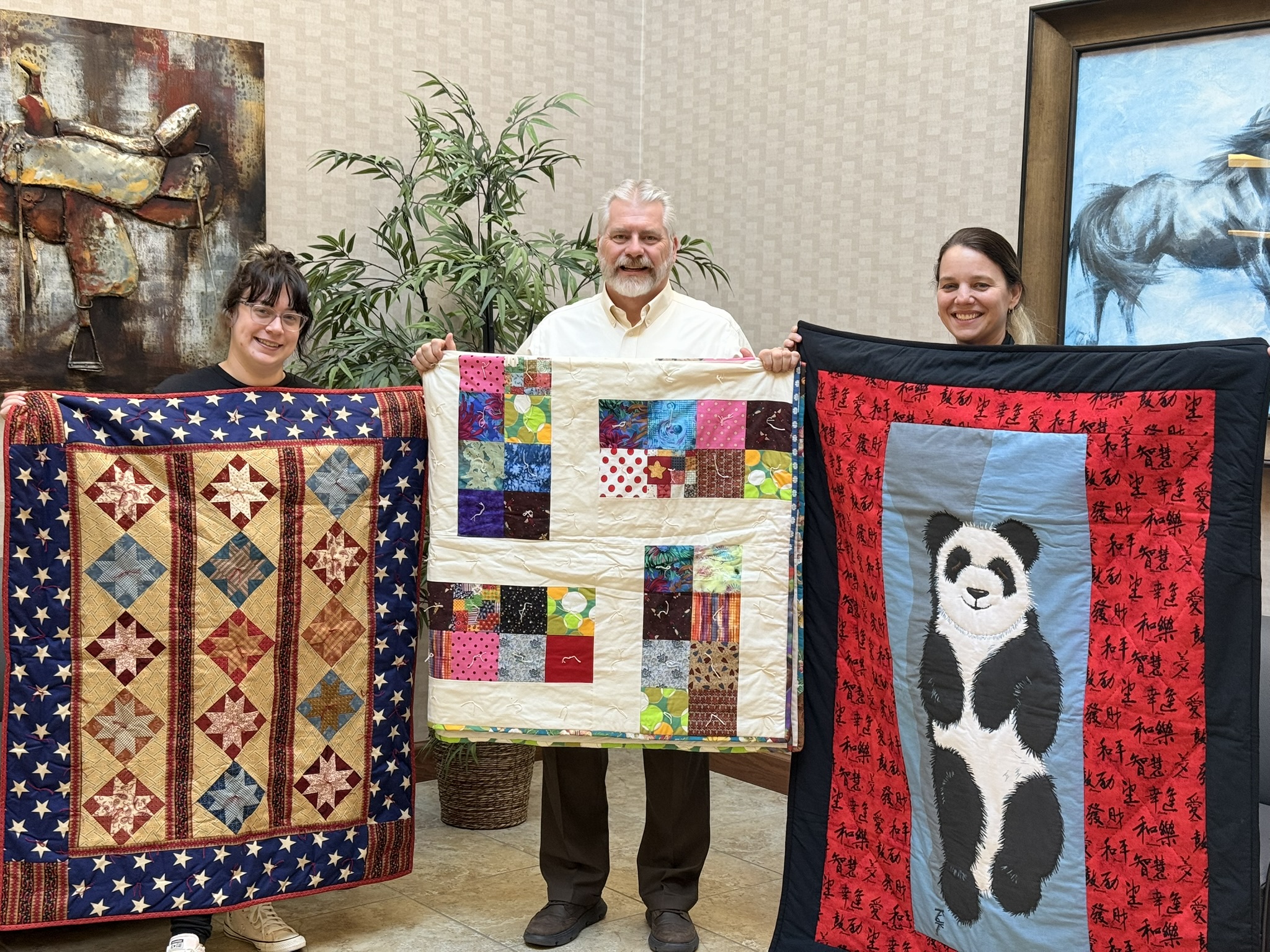 Picture of ASB President/CEO, Dave Hanson, and Marie Collins, Executive Director, and staff person Ali Lass from the Family Crisis Shelter holding up the quilts being donated. 